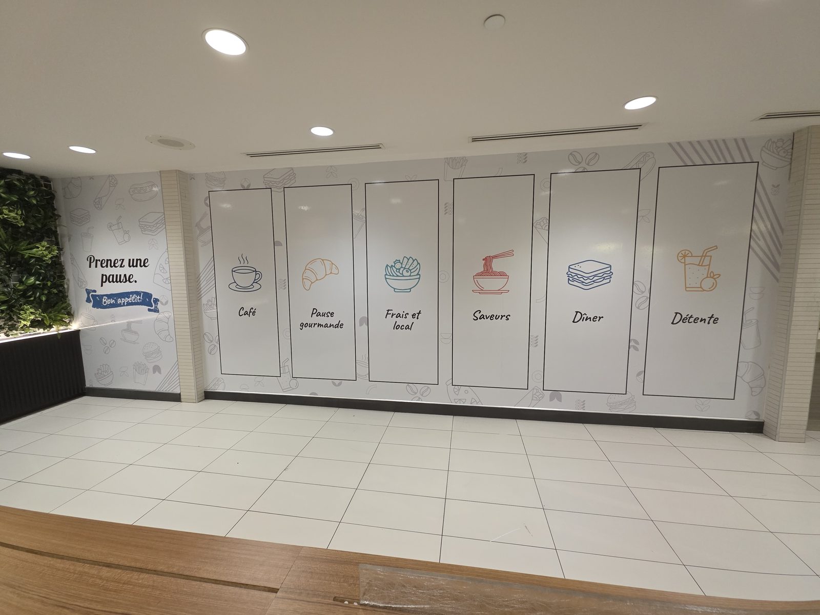 Food court feature wall covered in an illustrated food pattern with six framed panels labelled Café to Détente, continuing onto the side wall